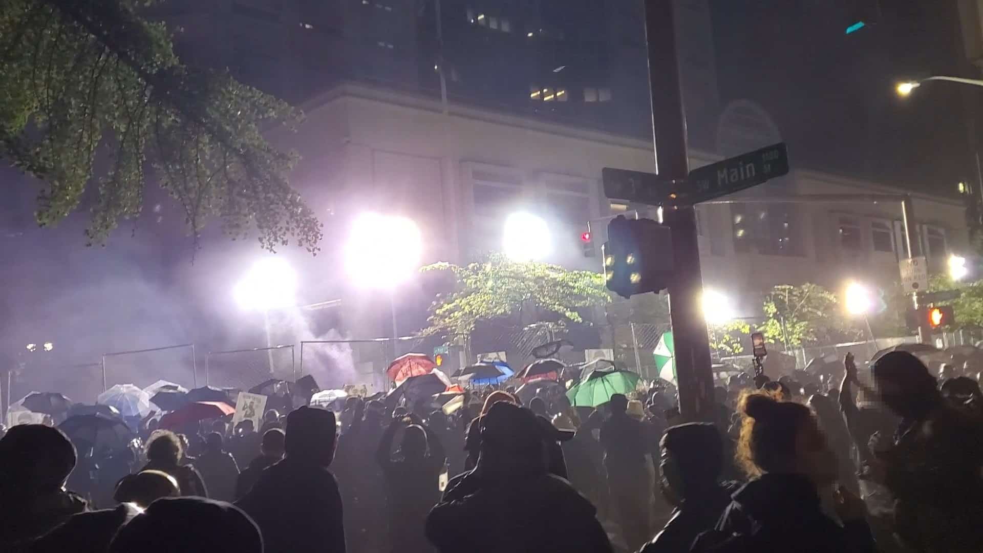 A crowd of protesters, many holding umbrellas, stand in front of the tall fencing surrounding the Justice Center at night. Blindingly bright lights shine down on them from the other side of the fence.