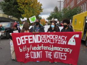 Five women march holding a large banner for the Defend Democracy coalition.