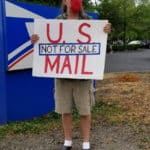A member of XRPDX stands next to a sign marking the entrance of a parking lot for a United States Post Office facility, holding a sign reading "U.S. MAIL: NOT FOR SALE"