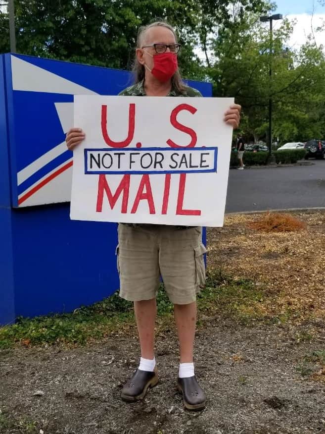 A member of XRPDX stands next to a sign marking the entrance of a parking lot for a United States Post Office facility, holding a sign reading "U.S. MAIL: NOT FOR SALE"