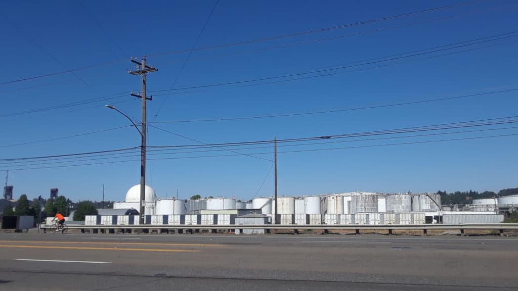 A photo from a distance of the Zenith Energy facility in Northwest Portland, comprised of a large number of white, largely nondescript cylinders and domes, with similarly bland train boxcars lined up in front.
