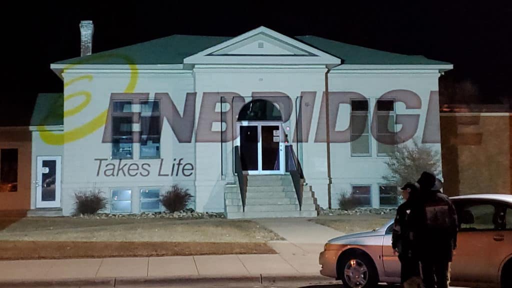 The Enbridge Energy office in Park Rapids, MN at night. A projector is covering the building with a slide showing the Enbridge logo, except with their slogan "Life Takes Energy" changed to say "Takes Life".