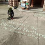 Outside of Portland City Hall, a member of XRPDX wearing all manners of flowers kneels next to messages written in colorful chalk: "NO ZENITH BOMB TRAINS" and "NO TAR SANDS Oil Trains In Portland or anywhere Else!"