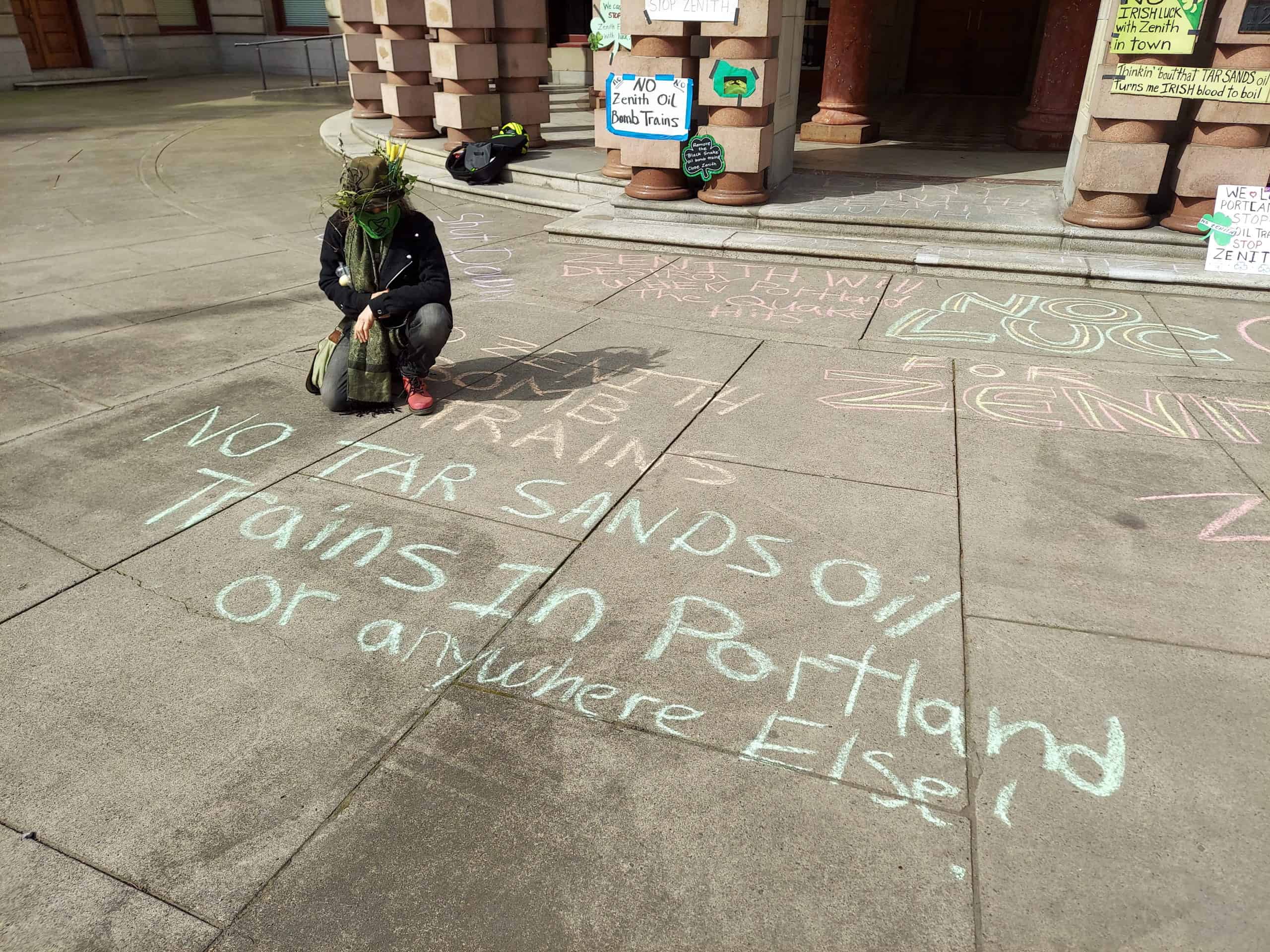 Outside of Portland City Hall, a member of XRPDX wearing all manners of flowers kneels next to messages written in colorful chalk: "NO ZENITH BOMB TRAINS" and "NO TAR SANDS Oil Trains In Portland or anywhere Else!"