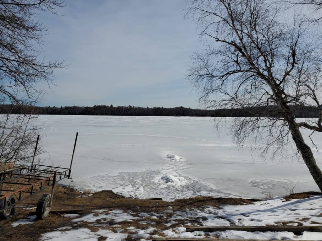 A photo of a frozen Minnesota lake, covered in snow. Some footprints have gone out a few meters onto the ice. The pathway is framed by a bare tree on the right and an empty, forgotten boat trailer on the left.