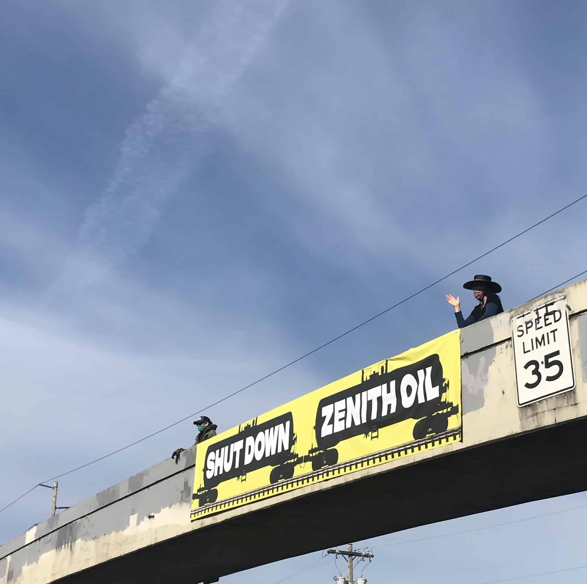 Two people standing on a pedestrian overpass holding a yellow banner that says "Shutdown Zenith Oil".