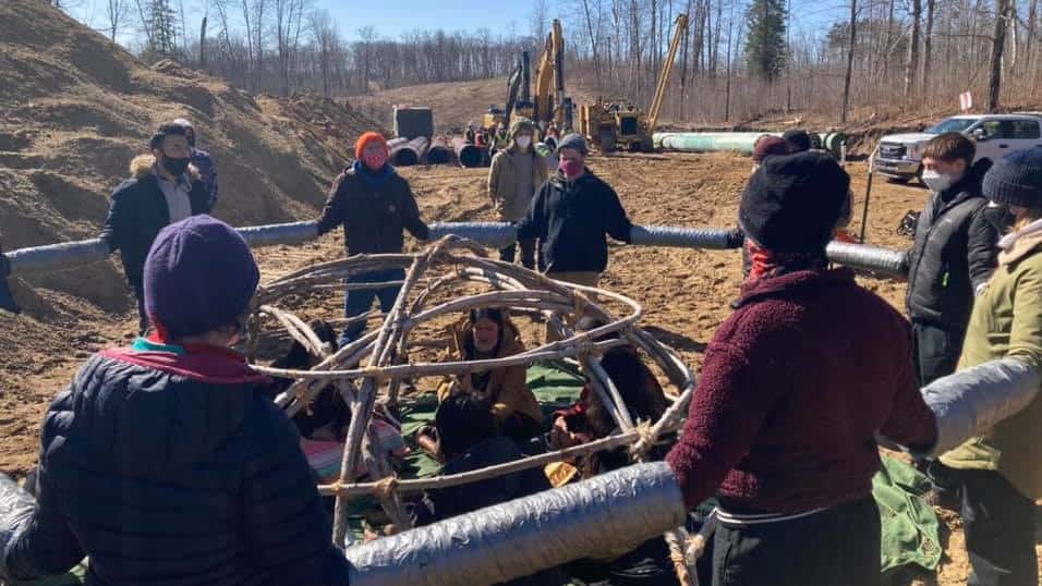In an Enbridge worksite, all dirt and heavy machinery, a number of Indigenous activists sit in a prayer lodge--an open dome made of branches--as non-Indigenous allies stand in a circle surrounding them with their hands in tubes taped together to make a circle (a tactic to make it difficult to remove any of them).