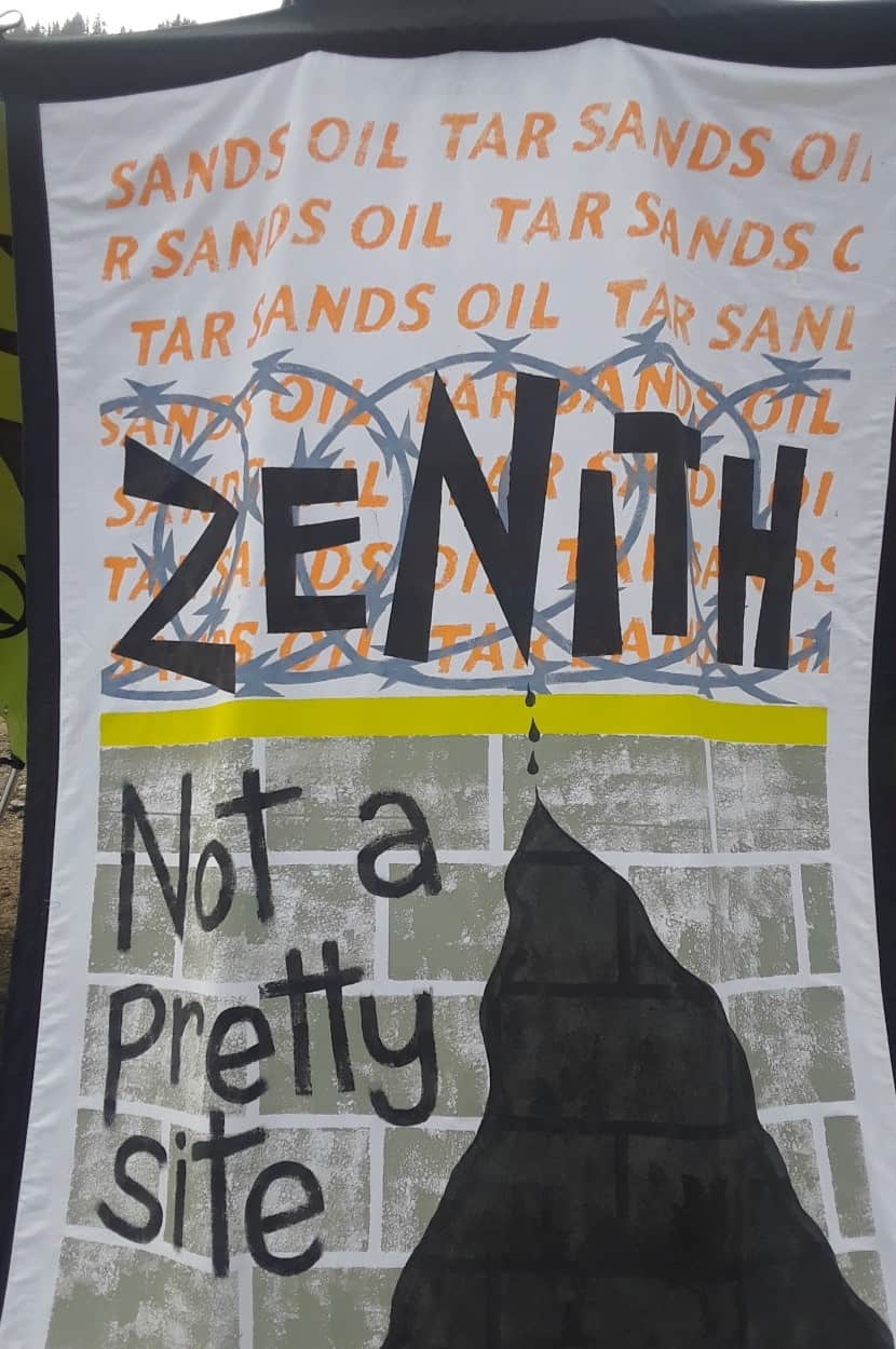 A screenprinted fabric that might be attached to a bag or jacket. It reads "ZENITH" over stylized barbed wire, above a grey brick wall which says "Note a pretty site." Oil drips down fron the N in Zenith to form a black mass down the wall. In the white space above the barbed wire, the words "TAR SANDS OIL" are written over and over in light orange.