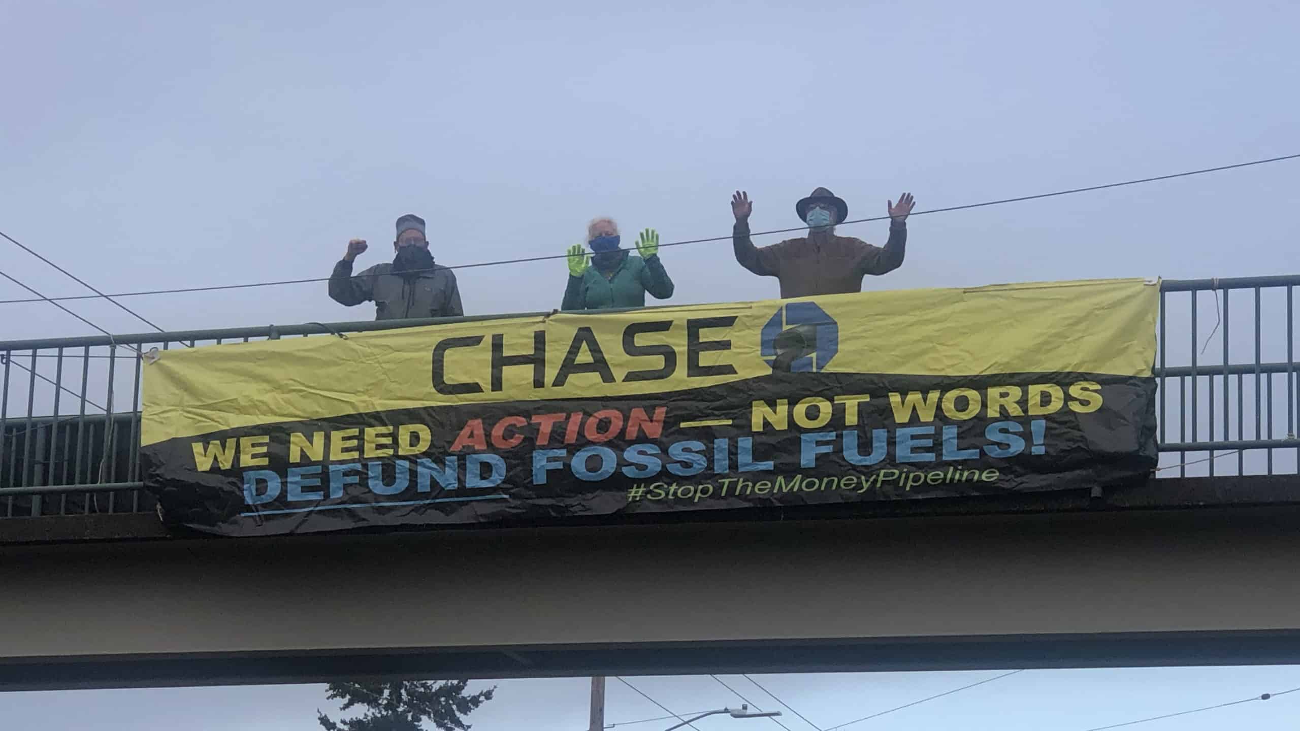 A photo from below of three people standing on a pedestrian overpass, waving their hands. Draped on the railing is a big yellow and black banner with the Chase Bank logo and, written below it: "WE NEED ACTIONS -- NOT WORDS. DEFUND FOSSIL FUELS! #StopTheMoneyPipeline"