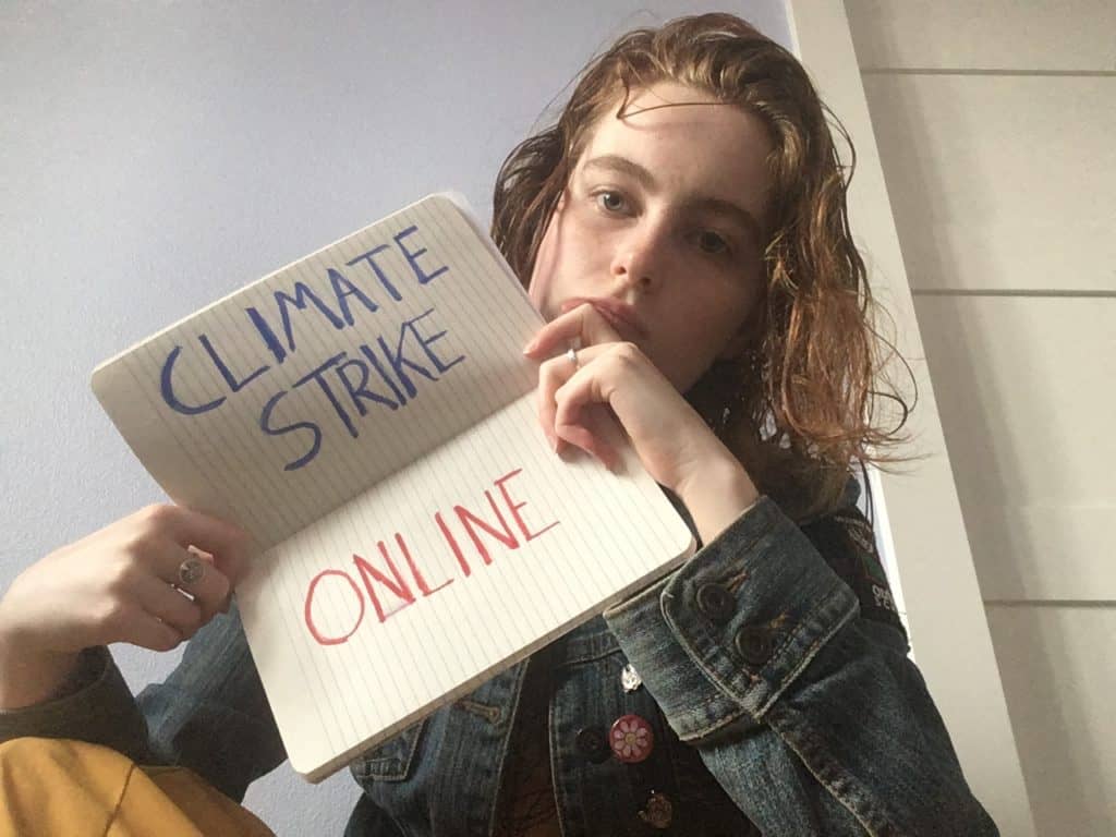 A young person with bright red hair and a blue denim jacket sitting in a room holds up a notebook in which the words CLIMATE STRIKE ONLINE