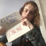 A young person with bright red hair and a blue denim jacket sitting in a room holds up a notebook in which the words CLIMATE STRIKE ONLINE