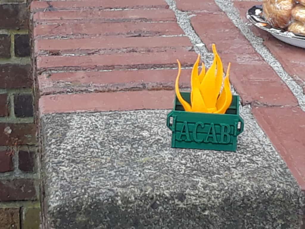 A picture of a green box sitting on a brick sidewalk with fake flames coming out of it. On the side is the acronym ACAB.