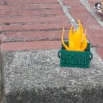 A picture of a green box sitting on a brick sidewalk with fake flames coming out of it. On the side is the acronym ACAB.