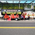 A view of the Tank of Doom--an oil car mock up used at protests--out in front of the Zenith Energy facility with protesters around it holding fake flames and sign saying "Stop Zenith Oil!"