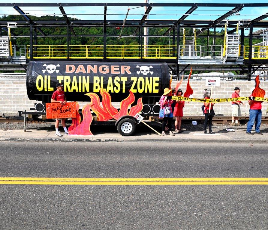 A view of the Tank of Doom--an oil car mock up used at protests--out in front of the Zenith Energy facility with protesters around it holding fake flames and sign saying "Stop Zenith Oil!"