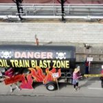 An areal view of the Tank of Doom--an oil car mock up used at protests--out in front of the Zenith Energy facility with protesters around it holding fake flames and sign saying "Stop Zenith Oil!"