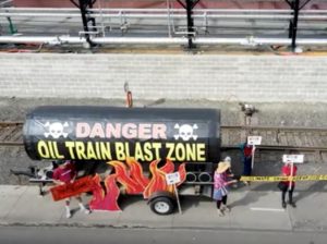 An areal view of the Tank of Doom--an oil car mock up used at protests--out in front of the Zenith Energy facility with protesters around it holding fake flames and sign saying "Stop Zenith Oil!"