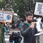 Man on microphone in front of 2 signs about climate justice